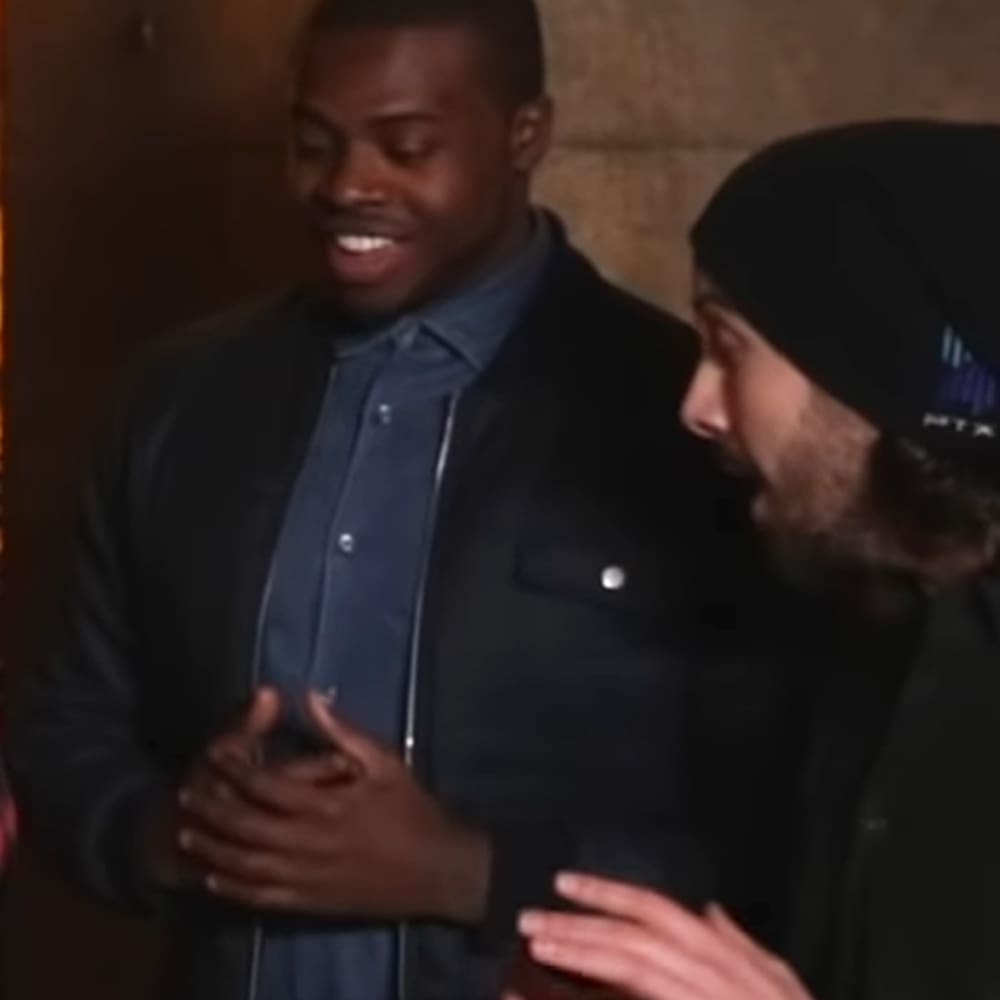 Young adult male in a dark jacket with light-colored buttons and pockets, standing indoors near another adult male wearing a black beanie and dark jacket, neutral-colored wall with even artificial lighting, indoor setting possibly an office or casual meeting space.