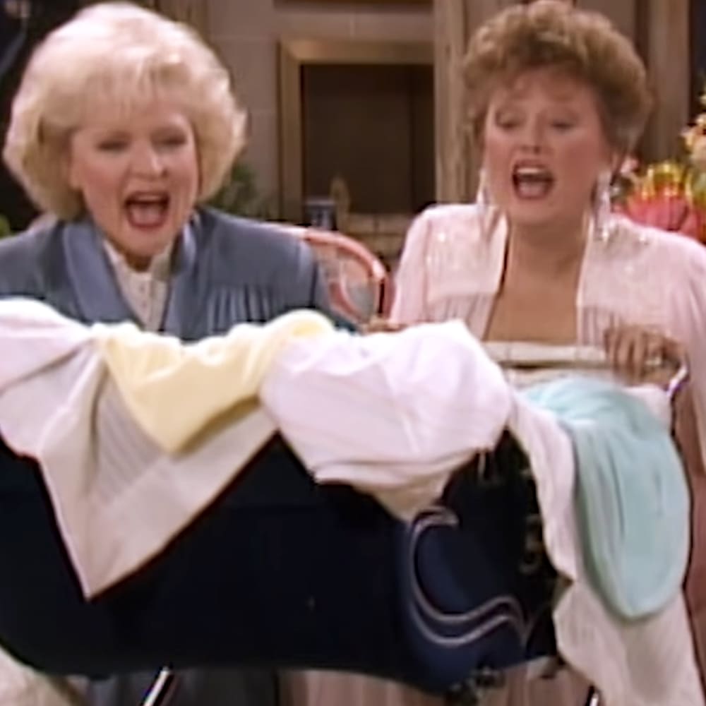 The Golden Girls' living room scene with two middle-aged females in a moment of surprise or shock; stack of folded clothes on a table, wooden furniture, natural light suggesting daytime, spring or summer ambiance.