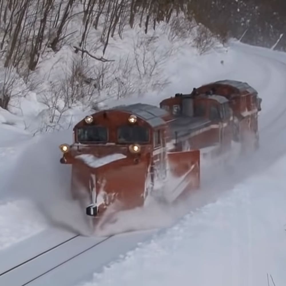 A red train moves through a snowy forest, clearing snow with its front plow in daylight.