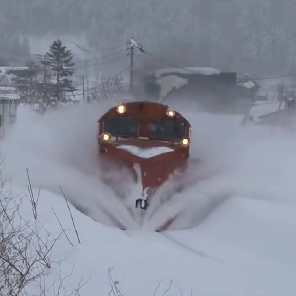 A red snowplow with headlights on clears a snowy rural road surrounded by snow-covered trees, power lines, and small buildings. The scene is set in winter at early morning or late afternoon.