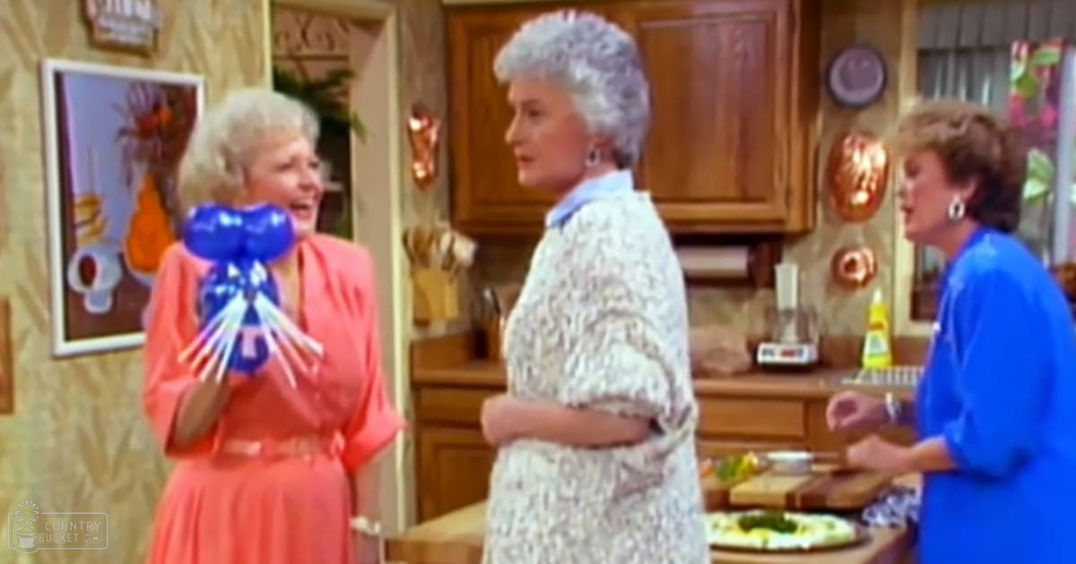 An adult female in an orange dress holds blue balloons with white ribbons, accompanied by two other adults in a kitchen setting.