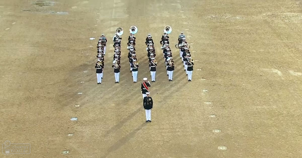 Young adult male band members performing outdoors on a sandy field under clear skies, dressed in dark uniforms with gold trim and light-colored pants.