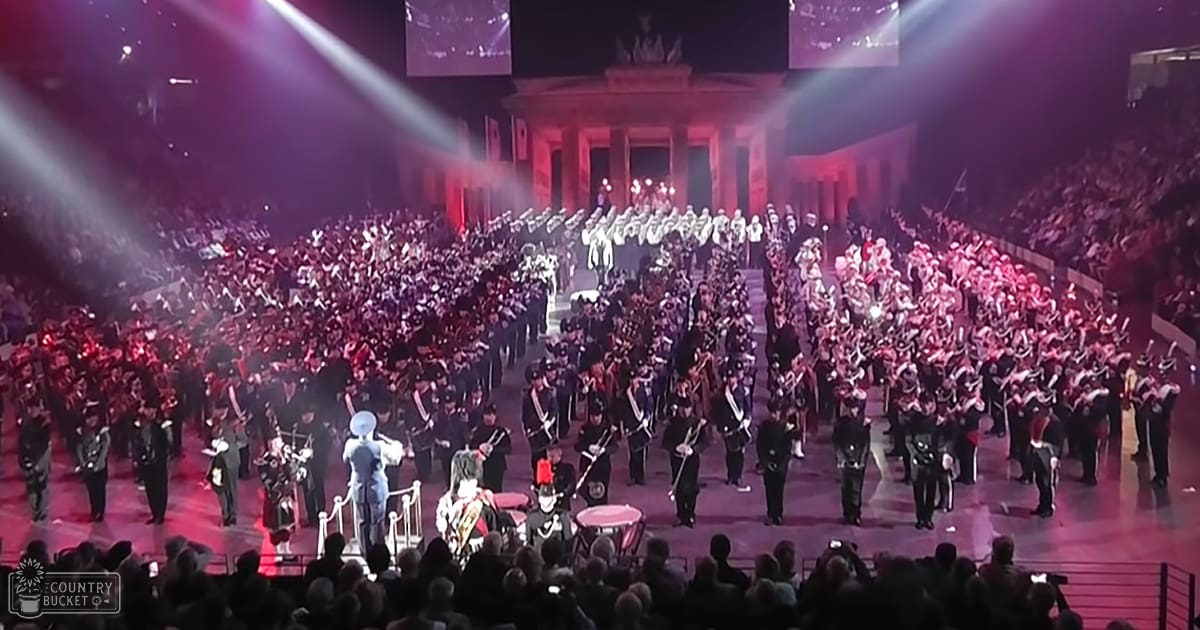 A large marching band performance in front of the Brandenburg Gate at night, with adults wearing formal military-style uniforms and a few female performers in elaborate costumes.