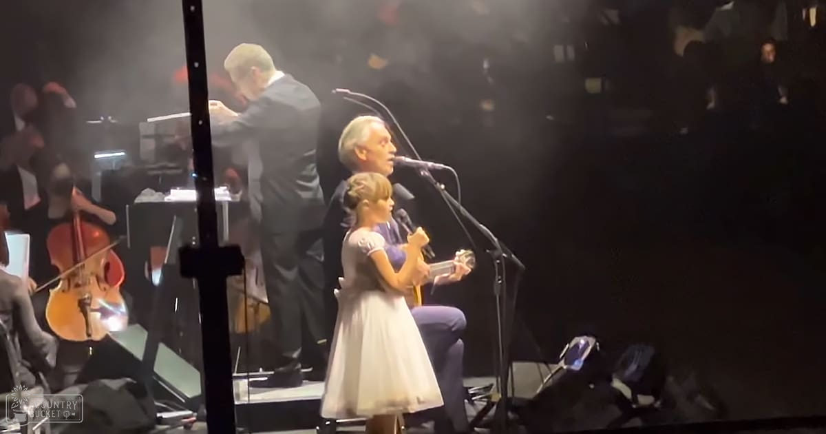 A young girl singing into a microphone at an indoor concert or performance setting, surrounded by adults in formal attire and musical instruments on stage.