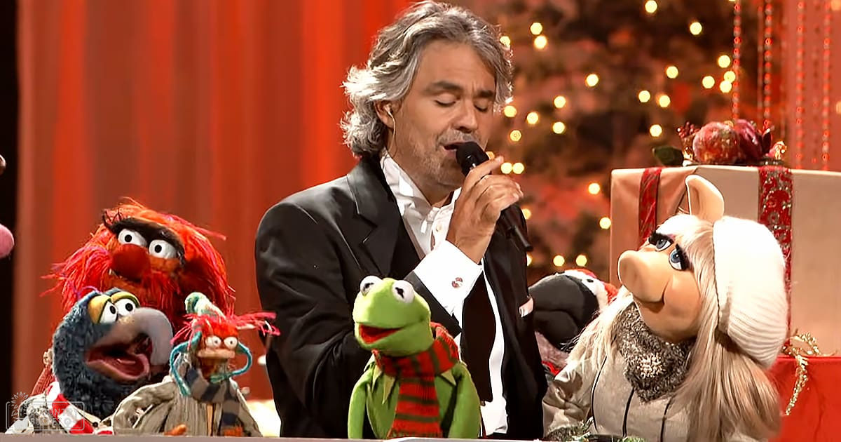 A festive indoor setting with Christmas decorations, including a tree adorned with lights and ornaments, wrapped presents, and a red curtain. An adult male singer is singing into a microphone surrounded by Muppets characters.