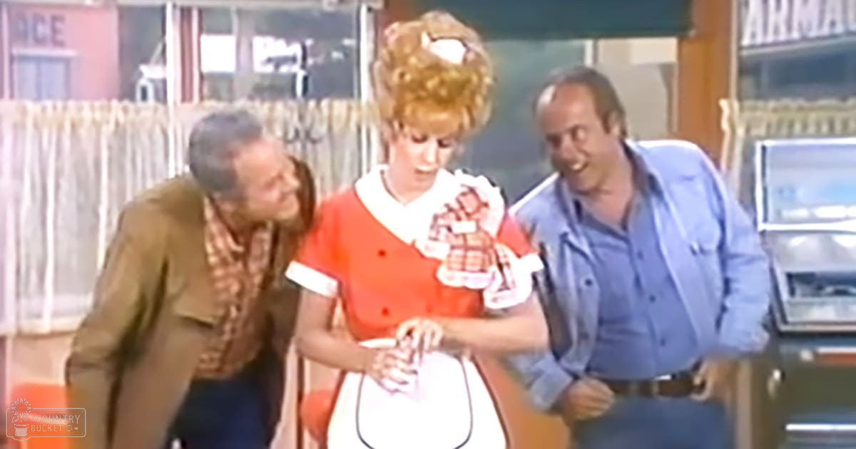 A woman in an orange dress with a white collar stands inside a store, holding what appears to be a white hat or bag. A middle-aged man wearing a brown jacket and plaid shirt stands nearby.