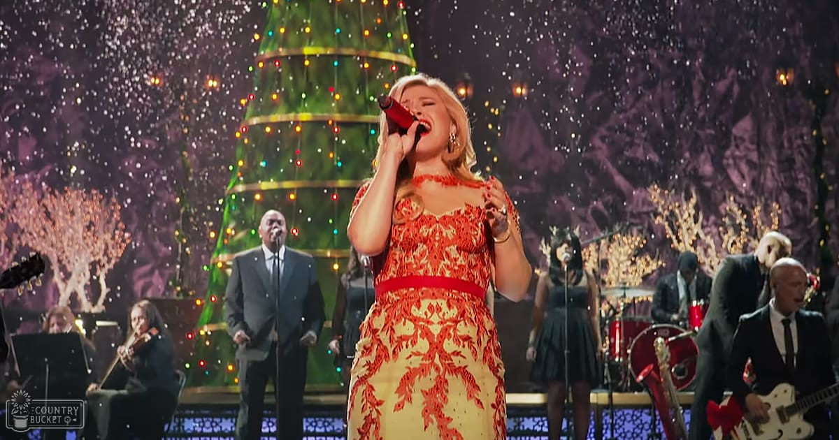 A woman singing in a festive Christmas-themed concert setting, wearing a red and gold patterned dress. Musicians in formal attire play instruments on stage with sheet music stands visible.