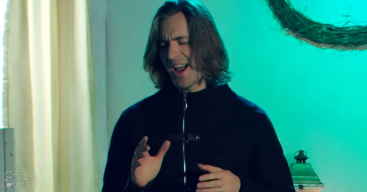 Young adult male wearing a dark blue zip-up jacket with a high collar, relaxed fit, and solid color. Indoor setting includes curtains, lampshade, and a branch or twig.