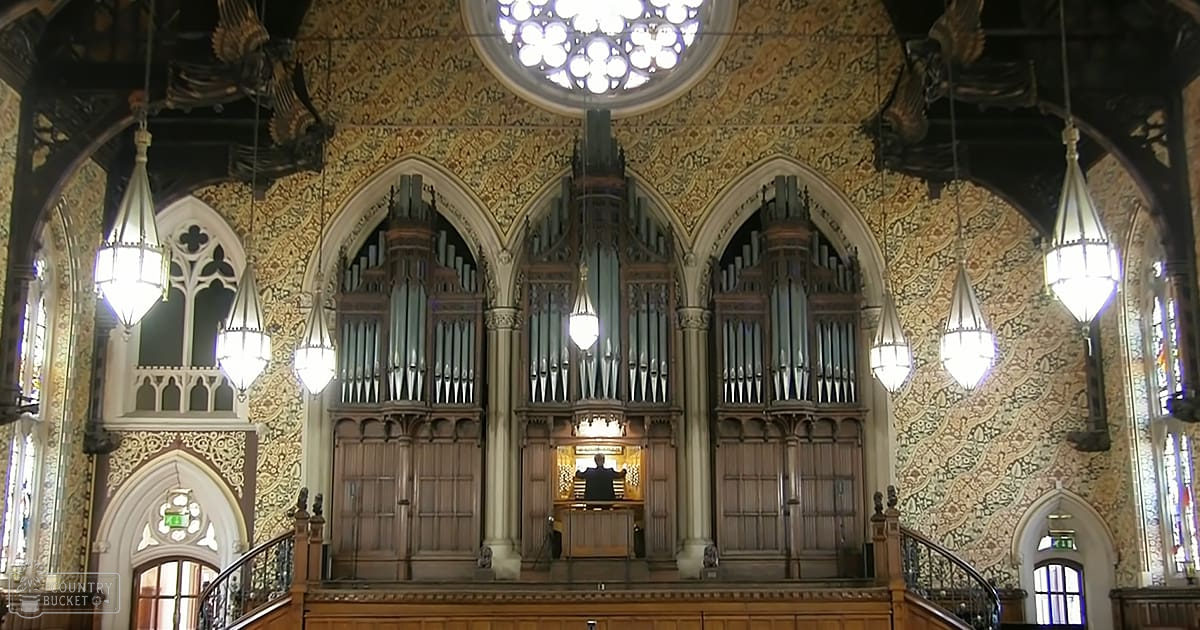 Adult male playing an ornate pipe organ in the interior of an ornately decorated church with high ceilings, detailed wall decorations, and stained glass windows on a sunny day.