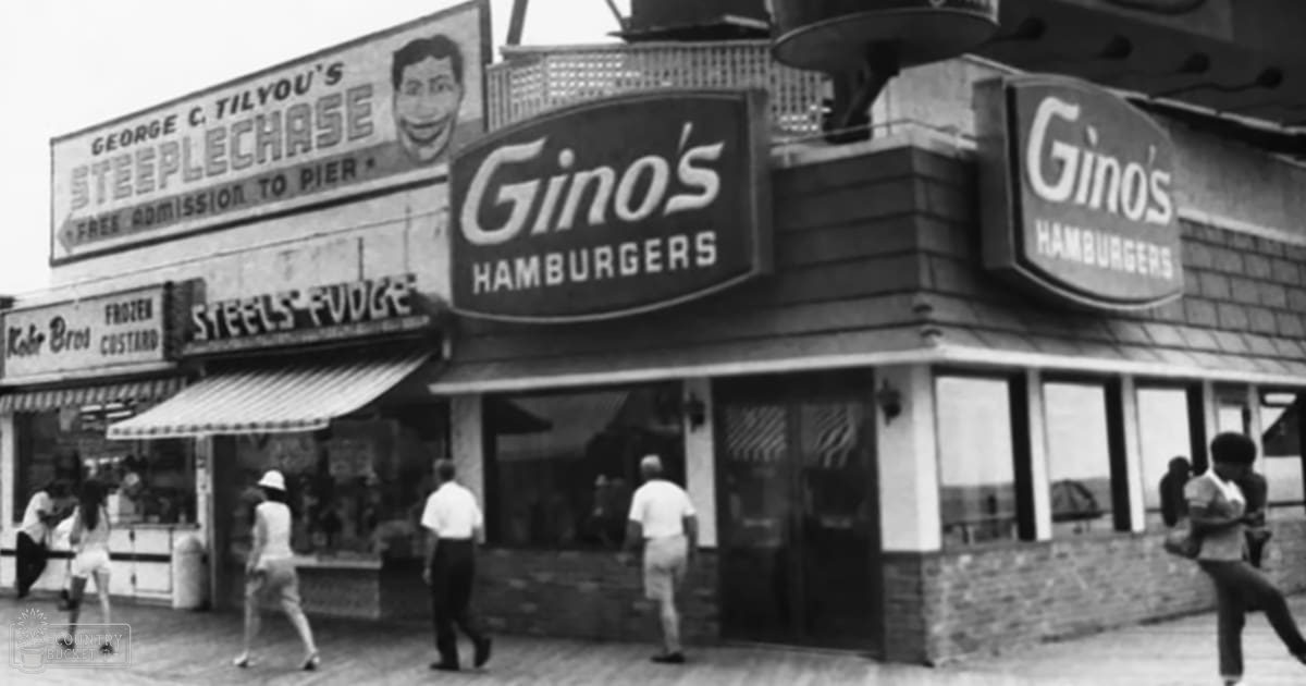An outdoor scene at a seaside amusement area with rides and food stalls in summer, with signs for 'George C. Tilyou's Steeplechase,' 'Kobr Bros Frozen Custard,' and 'Gino's Hamburgers' as people walk on a wooden boardwalk.