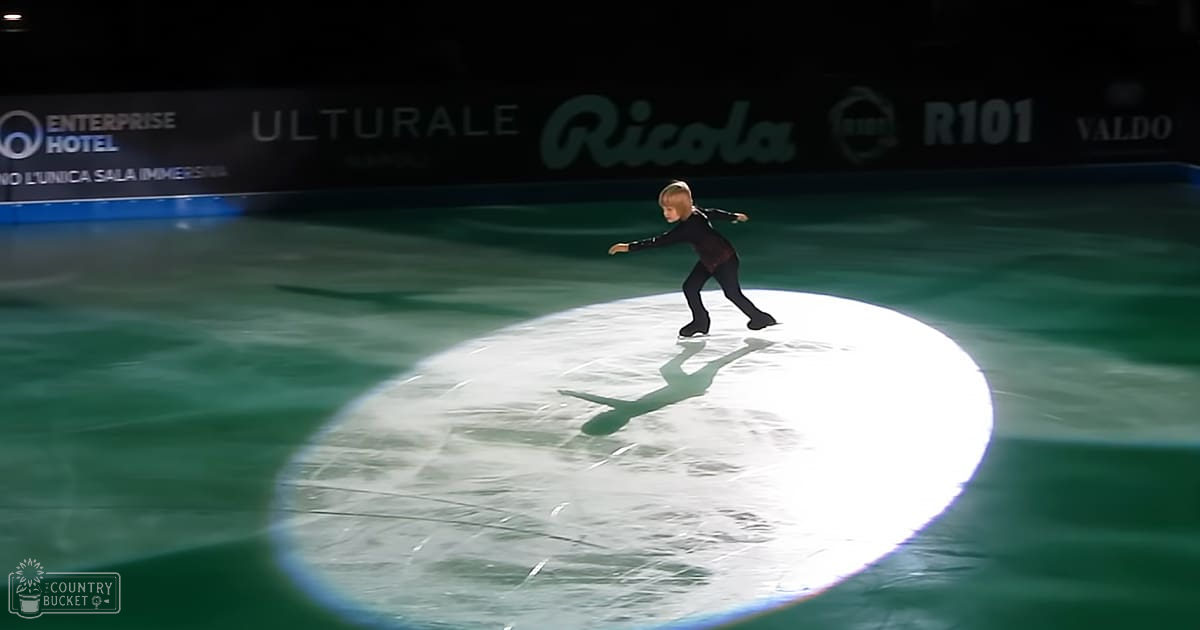 A young female figure skater in a black synthetic skating outfit performs on an indoor ice rink with green perimeter lighting.