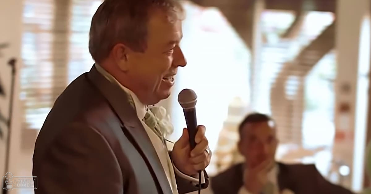A middle-aged man wearing a light-colored dress shirt with a greenish-yellow boutonniere speaks into a microphone at what appears to be an indoor wedding reception, while another young adult male in formal attire stands nearby.