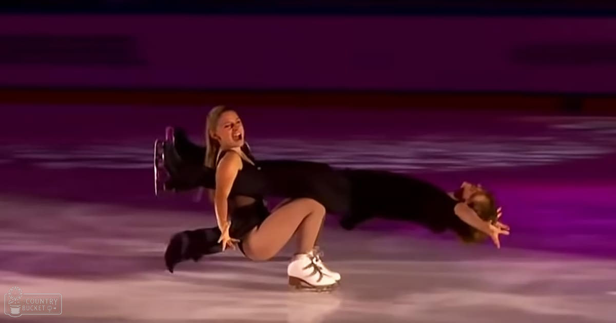 Young adult female figure skaters in a modern black figure skating outfit and white figure skates, performing on an indoor ice rink with purple lighting.
