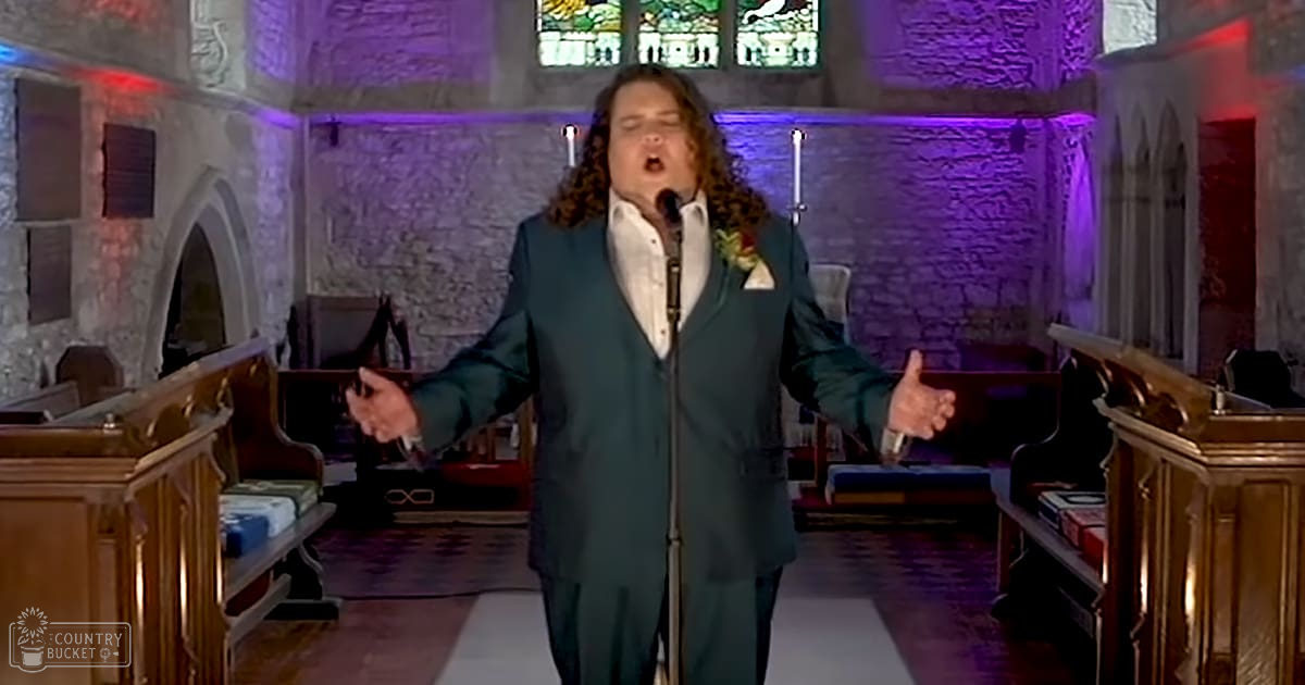 An adult male singer performing inside a church with wooden pews, stained glass windows, and decorative lighting on stone walls.