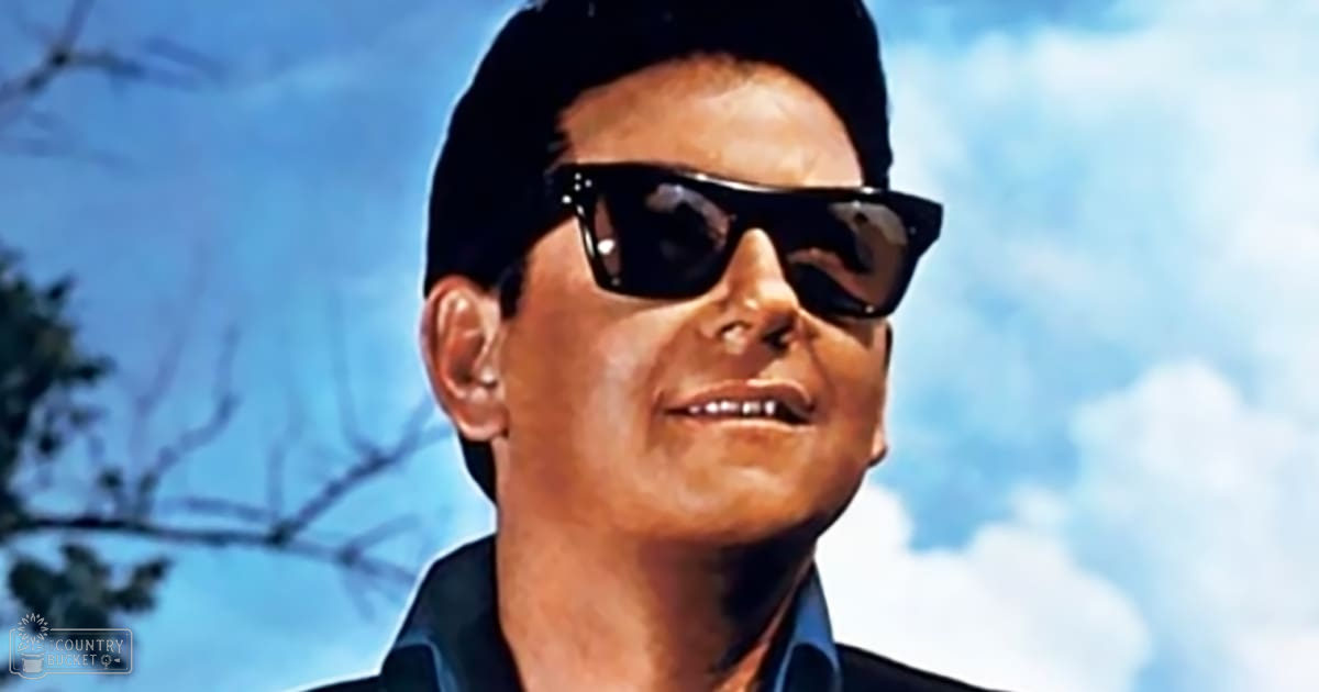Adult male in a relaxed, casual button-down shirt with long sleeves, wearing dark sunglasses and a cap, outdoors under a bright blue sky with scattered white clouds.