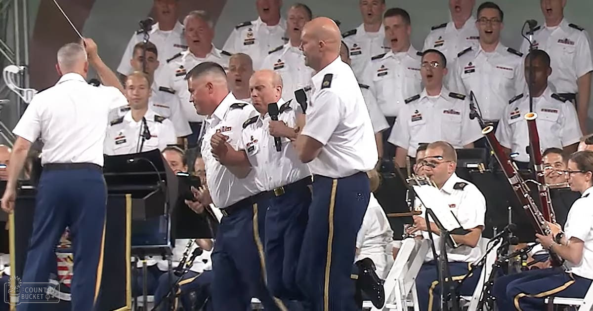 A military choir performing in an indoor concert hall with adults wearing white shirts with epaulets and dark blue pants with gold stripes, holding conductors baton and microphones. The setting is indoors, likely a stage area with neutral dark background.