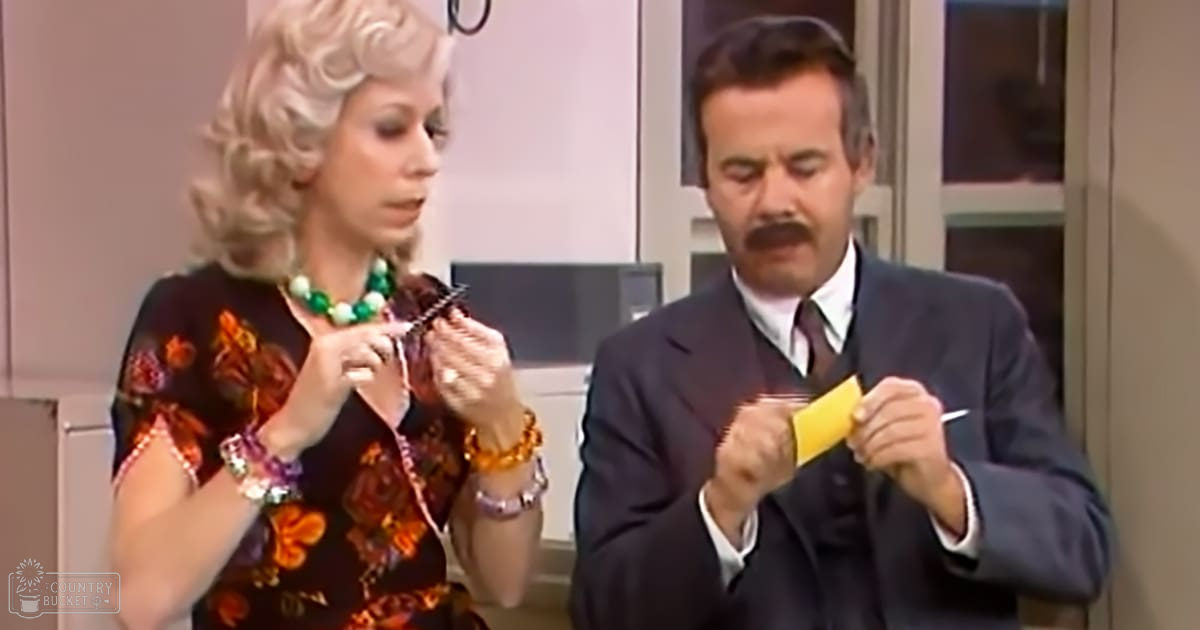 A comedic scene with two adults, one wearing a black dress with orange-red floral patterns and another in a dark suit jacket over a white shirt and tie, set indoors possibly in a living room or studio.