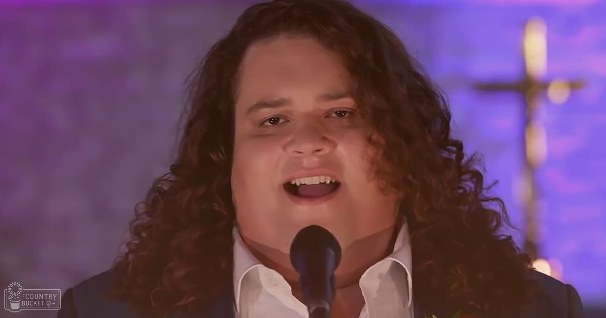 A young adult male singer in a formal shirt and jacket, singing into a microphone against religious imagery in an indoor setting with soft ambient lighting.