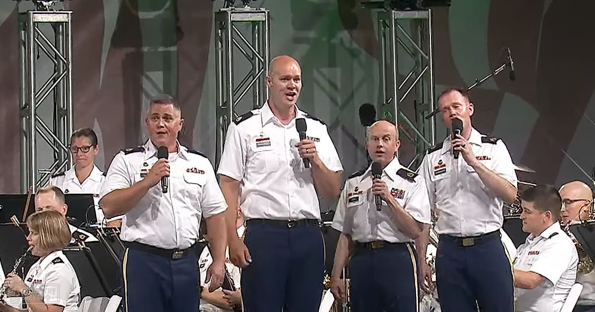 Group performance at an indoor event with performers in white shirts, dark blue pants, and black belts. Microphones and musical instruments are present on stage under dim lighting.