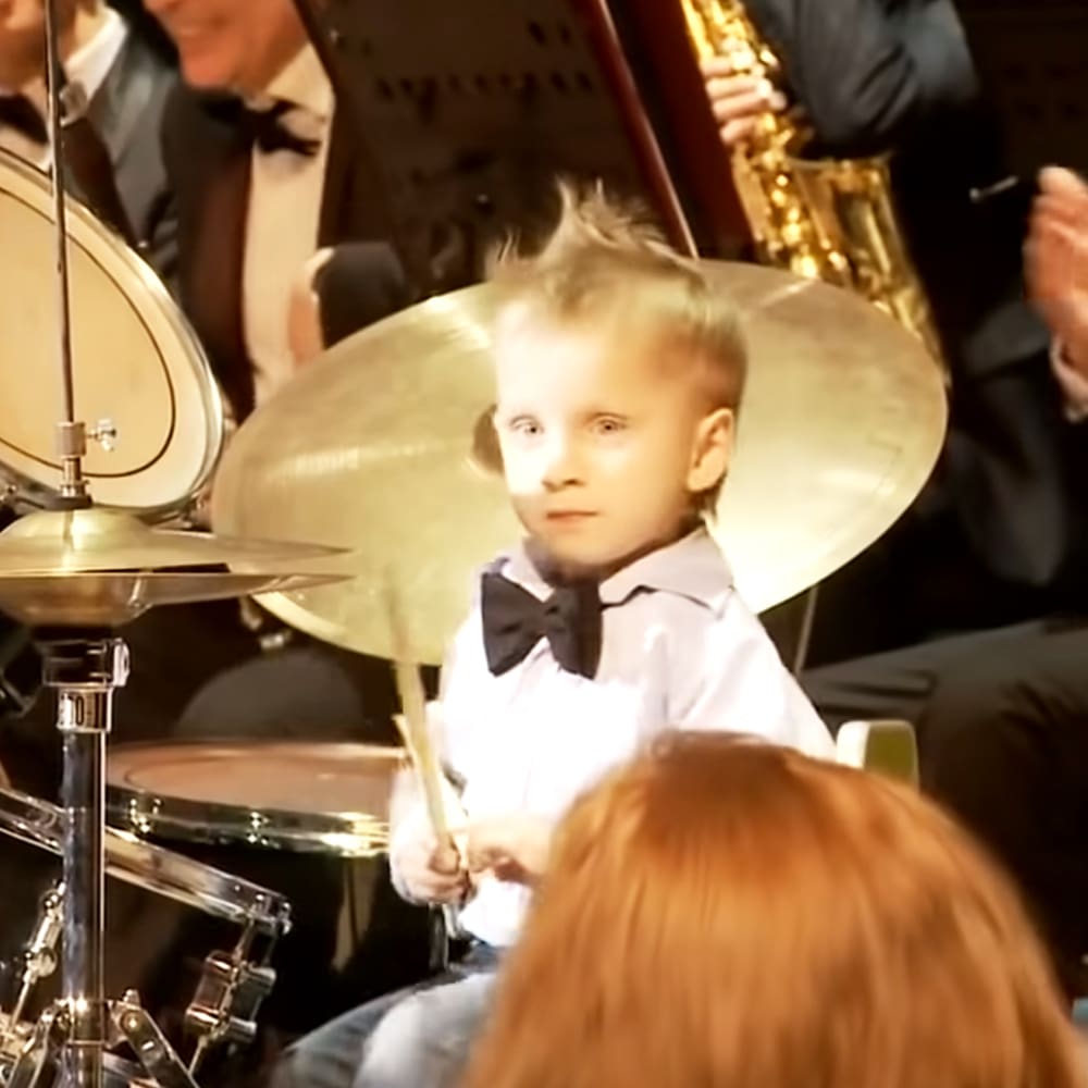 A young child plays drums at a formal concert setting with adults in suits and ties, with a saxophone and various musical instruments.