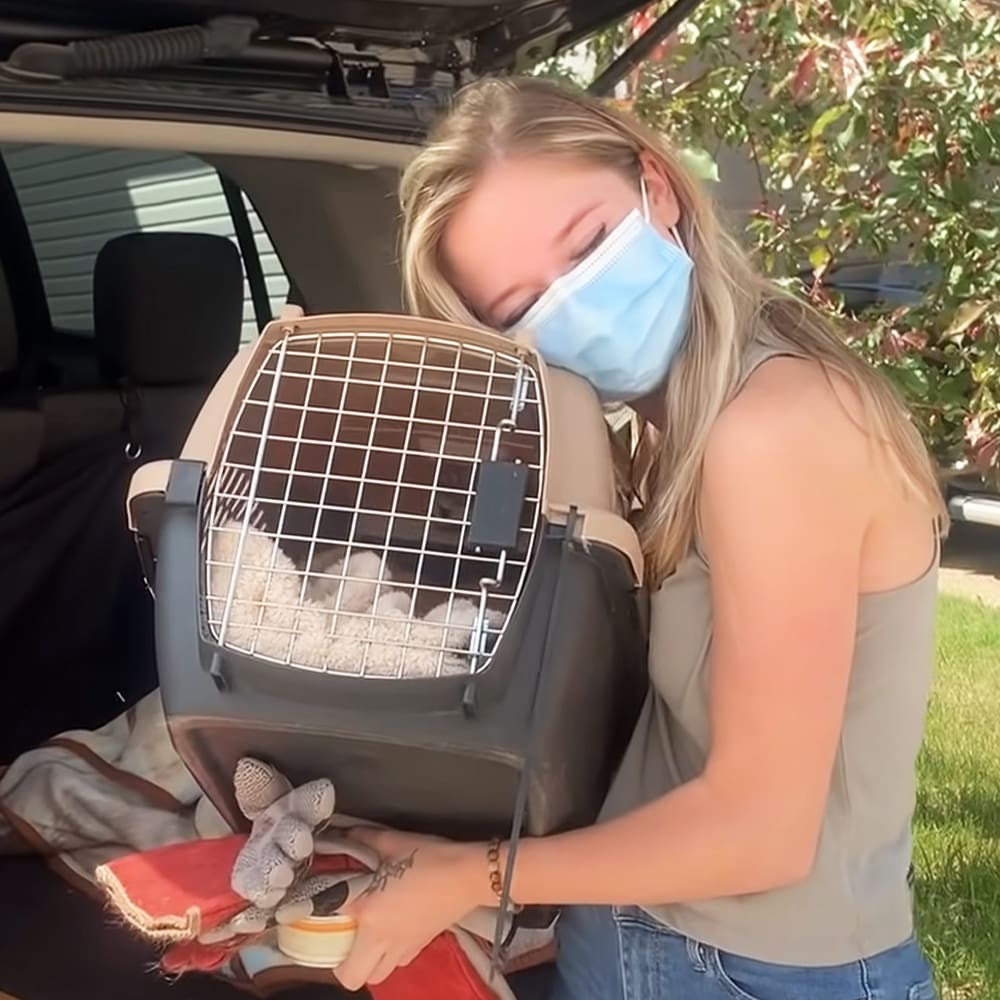 A young adult female wearing a gray sleeveless top and blue jeans, holding an animal carrier outdoors near an SUV with green grass and trees in the background.