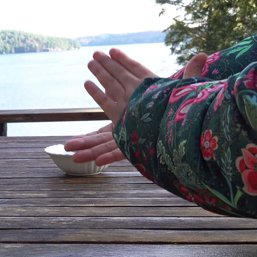 Adult female in floral-patterned loungewear sitting at an outdoor deck, hands extended towards a small white bowl on a wooden table with a body of water and greenery in the background.