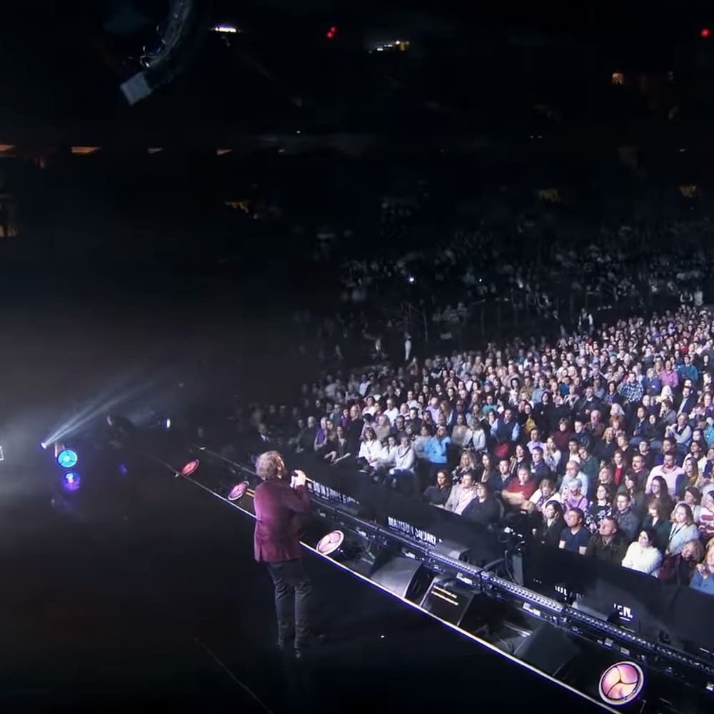 An adult male singer performs on stage in an indoor concert hall at night; audience members are seated in tiered rows wearing casual to semi-formal attire, illuminated by stage lights and ambient venue lighting.