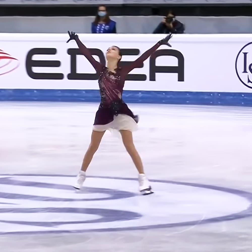 A young adult female figure skater in a maroon and black sequined leotard with white skirt, performing at an indoor ice rink.