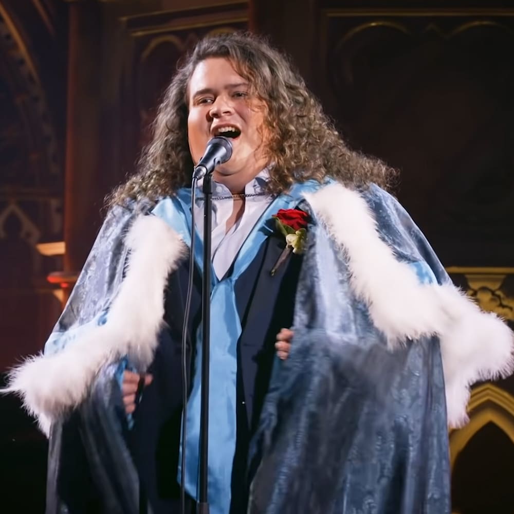 A young adult male in a blue velvet robe with white fur trim and a red rose boutonniere, performing behind a microphone stand within an ornate church or cathedral interior.