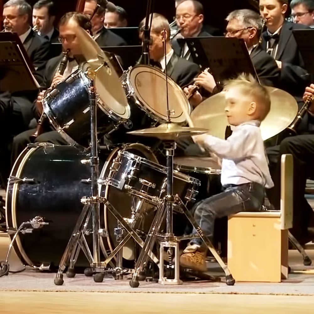 A toddler playing drums in an orchestra setting with adults in formal attire, daytime indoor venue with natural light and neutral tones.