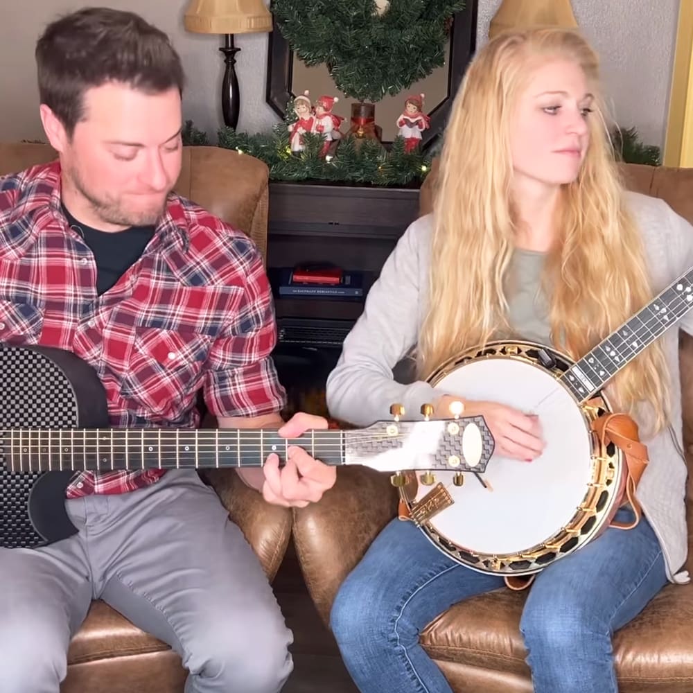 A young adult male singer in a red plaid shirt and gray pants performs alongside another young adult female wearing a light gray top and blue jeans, with a festive Christmas tree and decorations in the background.