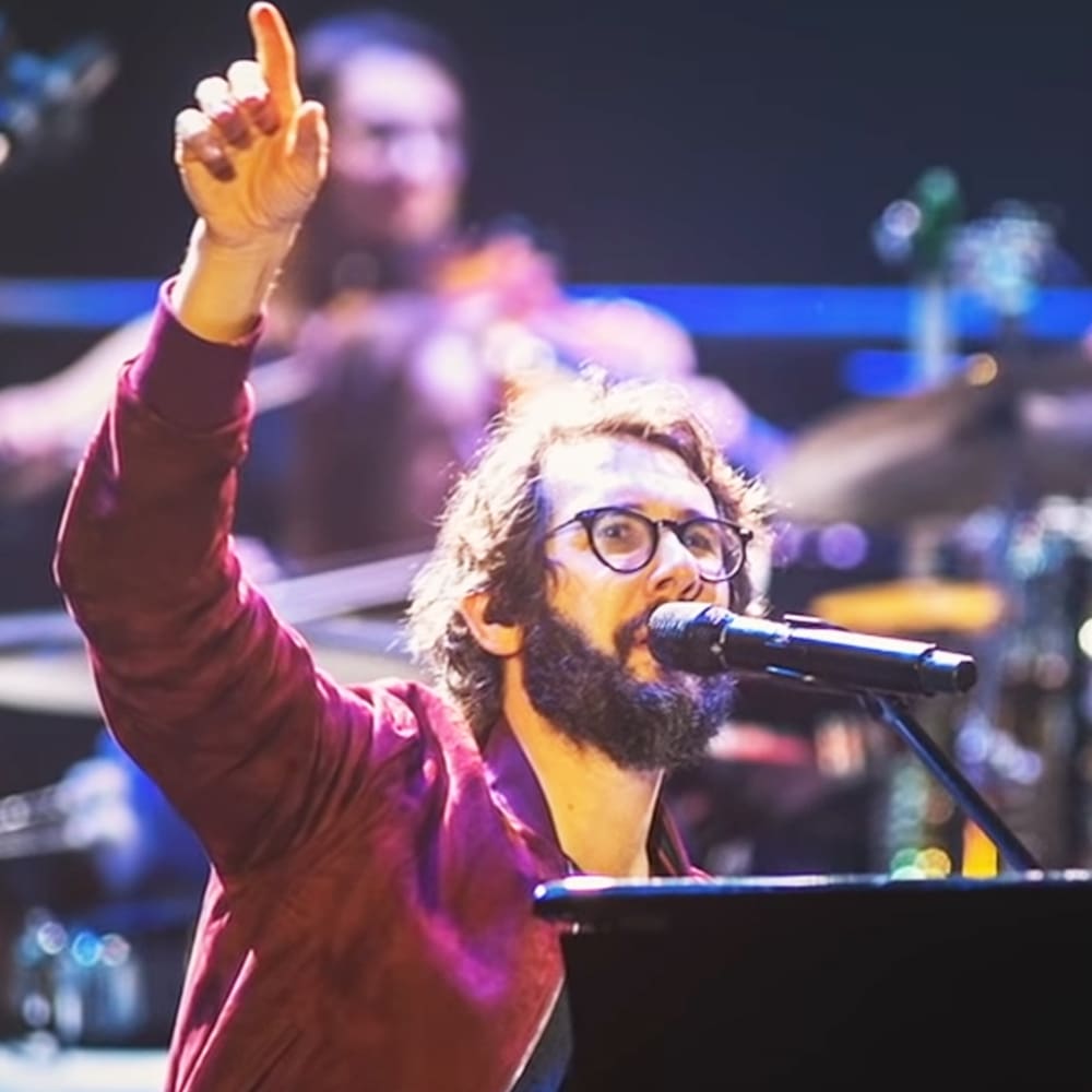 A male adult singer wearing a maroon long-sleeved shirt with rolled-up sleeves, performing at an indoor concert venue under stage lighting.