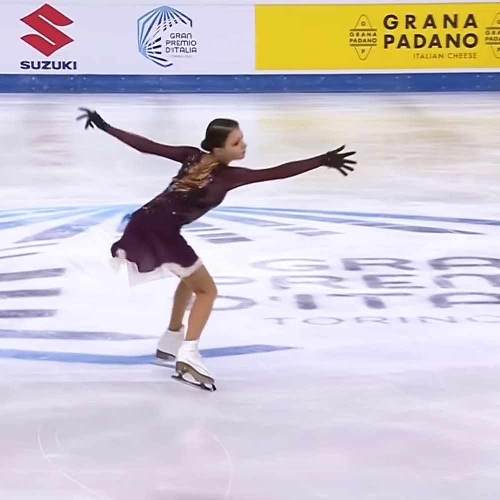 A young adult female figure skater performing at an indoor ice rink with artificial lighting, wearing a dark purple figure skating dress with long sleeves and a low-cut neckline.
