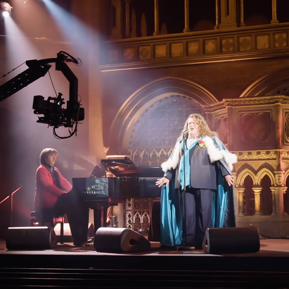 An adult female pianist in a red jacket and dark pants seated at a grand piano with ornate architecture, possibly within a historic church or cathedral; an older male performer stands on stage wearing a long coat with fur trim, blue suit, and sash under artificial evening light.