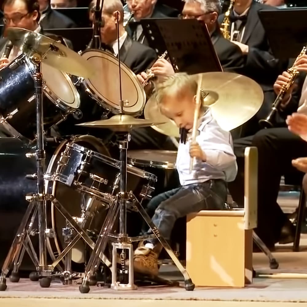 A young child plays drums on a small stool, surrounded by formally dressed adults seated behind him, in what appears to be an auditorium or concert hall setting under stage lighting.