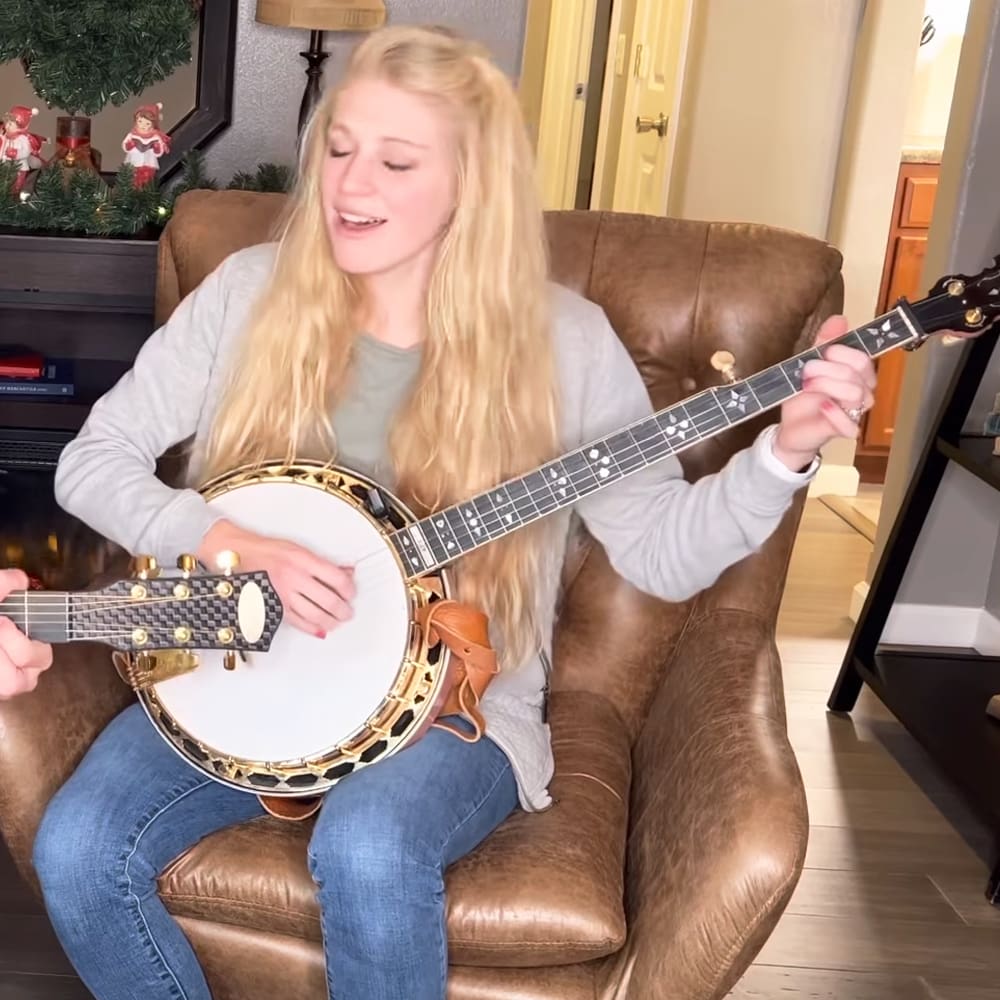 A young adult female playing a banjo in an indoor living room setting with Christmas decorations, natural daylight.
