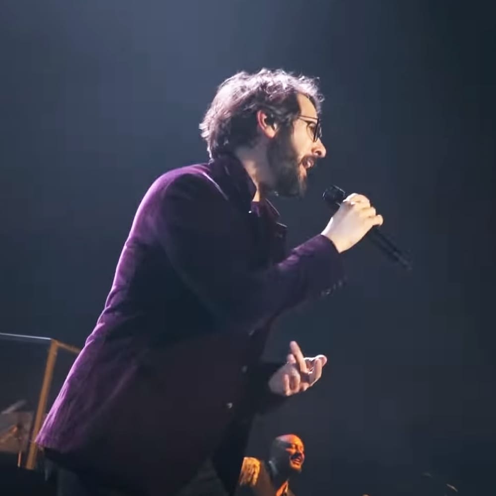 An adult male performer in a dark purple jacket and high-collared shirt, holding a microphone on stage during a concert or live event with another male performer in formal attire behind him under focused stage lighting.