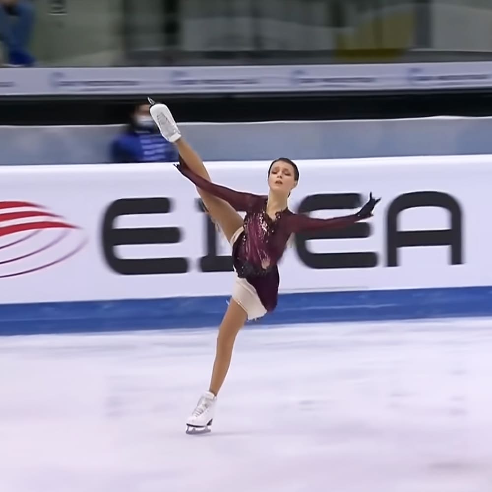 Young adult female figure skater in a maroon leotard with white skirt and black gloves, practicing indoors at an ice rink.