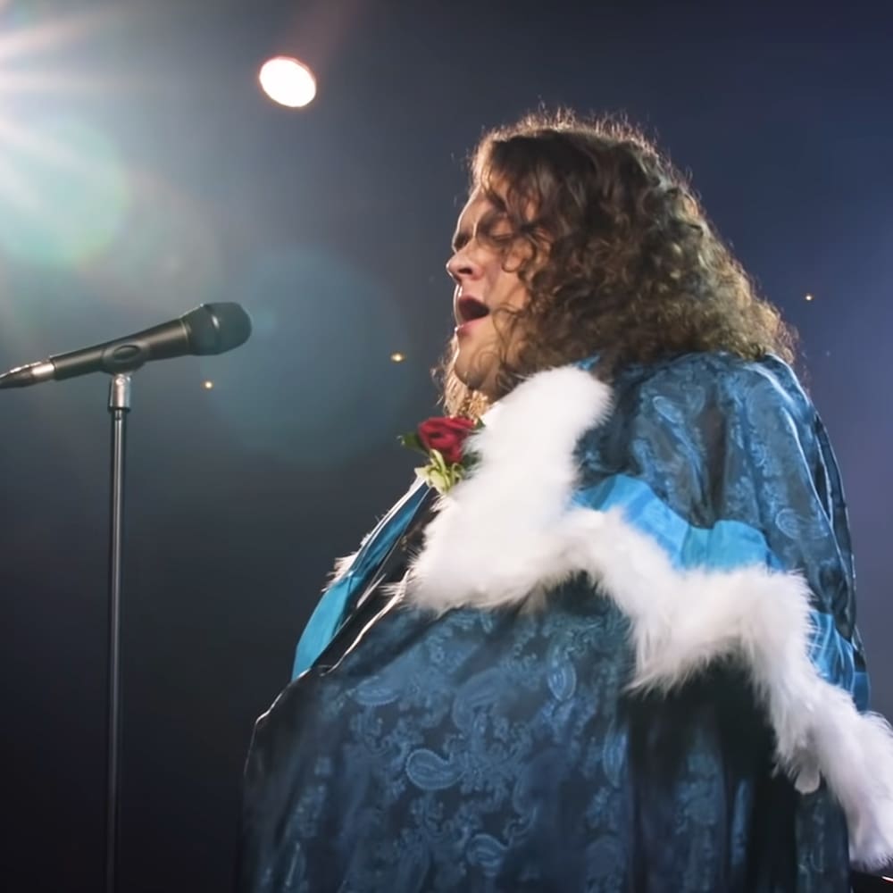 An adult male performer in a blue, intricately patterned garment with white fur trim and a red rose, positioned on an indoor stage with stage lighting flares at night.