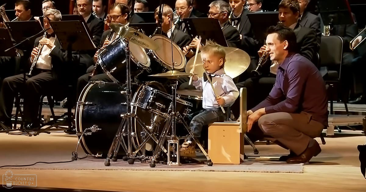 A young child plays drums in an indoor concert hall with sheet music stands and stage lighting, accompanied by a smiling adult male.