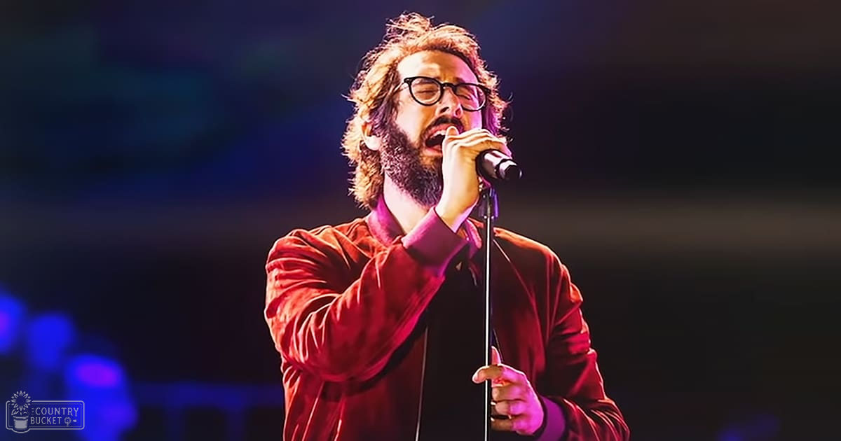 Adult male singer wearing a red velvet jacket at an indoor concert venue, holding a microphone with stage lighting and blurred background elements visible.