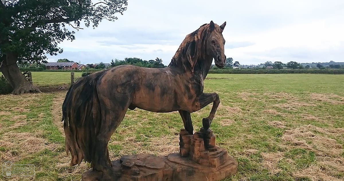 A horse statue in an open field with a tree on the left and houses in the background under spring or summer conditions.
