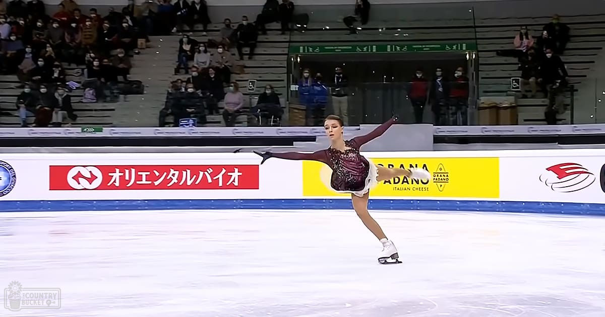 A young adult female figure skater performing at an indoor ice rink with a diverse audience wearing winter clothing. Advertisements for 'オリエンタルバイオ,' 'ORANA ADANO,' and 'ITALIAN CHEESE' are visible on the boards around the rink.