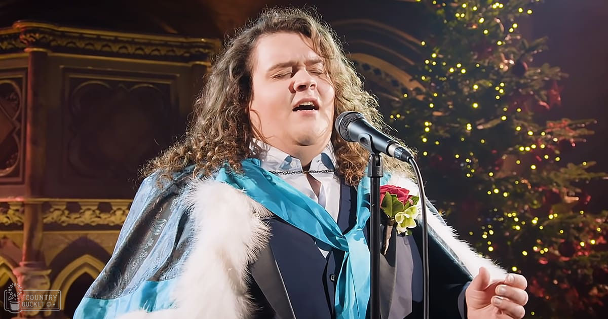 A male performer in a formal or festive outfit stands indoors, with an ornate background including a decorated Christmas tree and ornate architectural details.