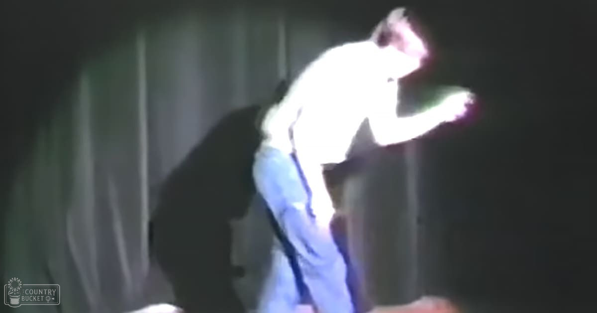A young child wearing light-colored jeans and a white shirt, standing indoors with plain wall in the background.