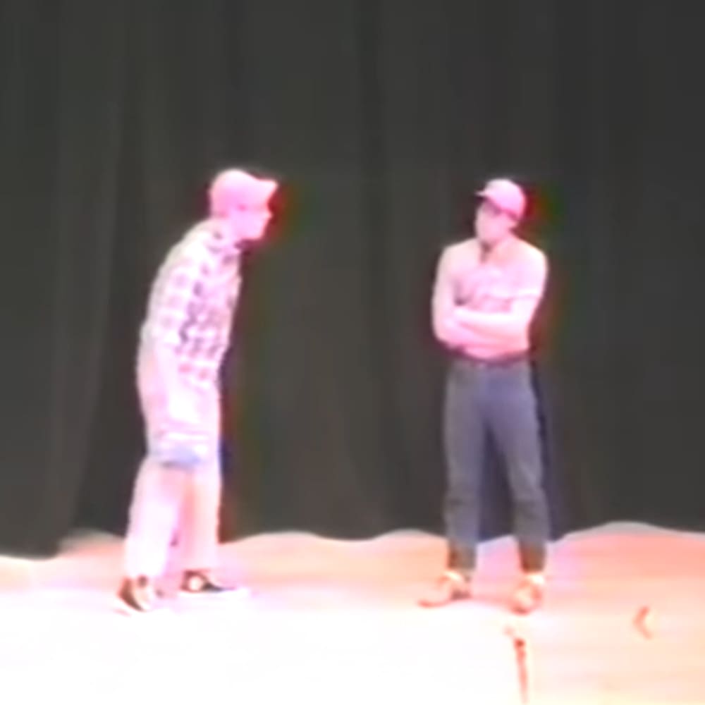 Two adult males dressed in casual attire perform on a stage with muted lighting.