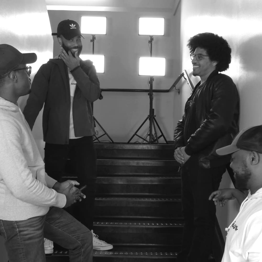 Group of four adult males, one holding something small, dressed casually with hoodies and sneakers, in a modern indoor studio setting with professional lighting equipment.