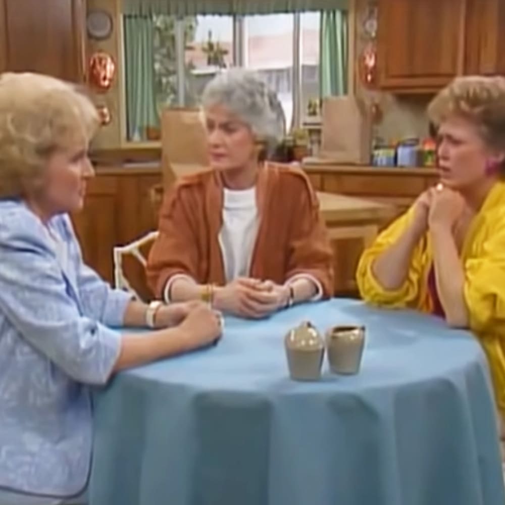 Three adult females in a kitchen setting with wooden cabinets and daylight through the window, wearing light blue button-up shirt, orange cardigan, and yellow blouse respectively.