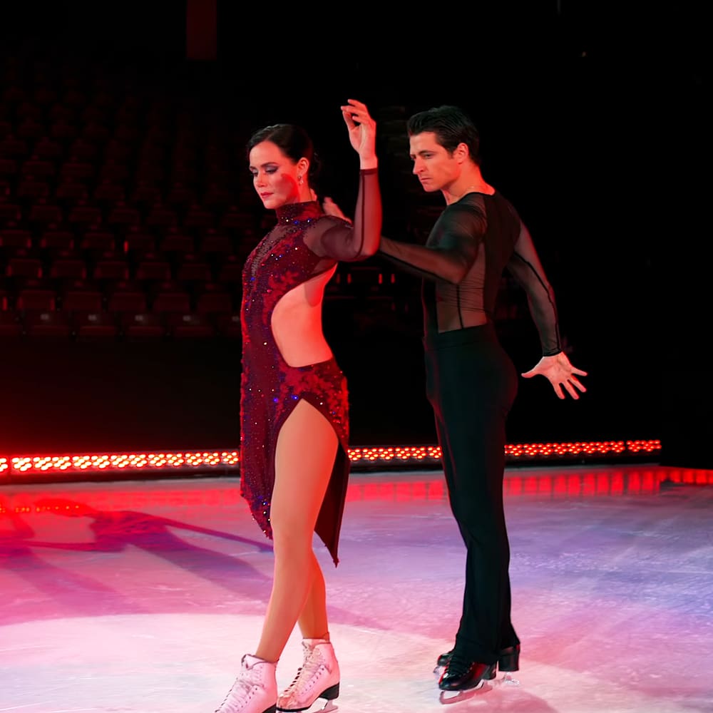 Figure skating pair performing on an ice rink in a dramatic indoor arena with red lighting during what appears to be practice or before an event begins.