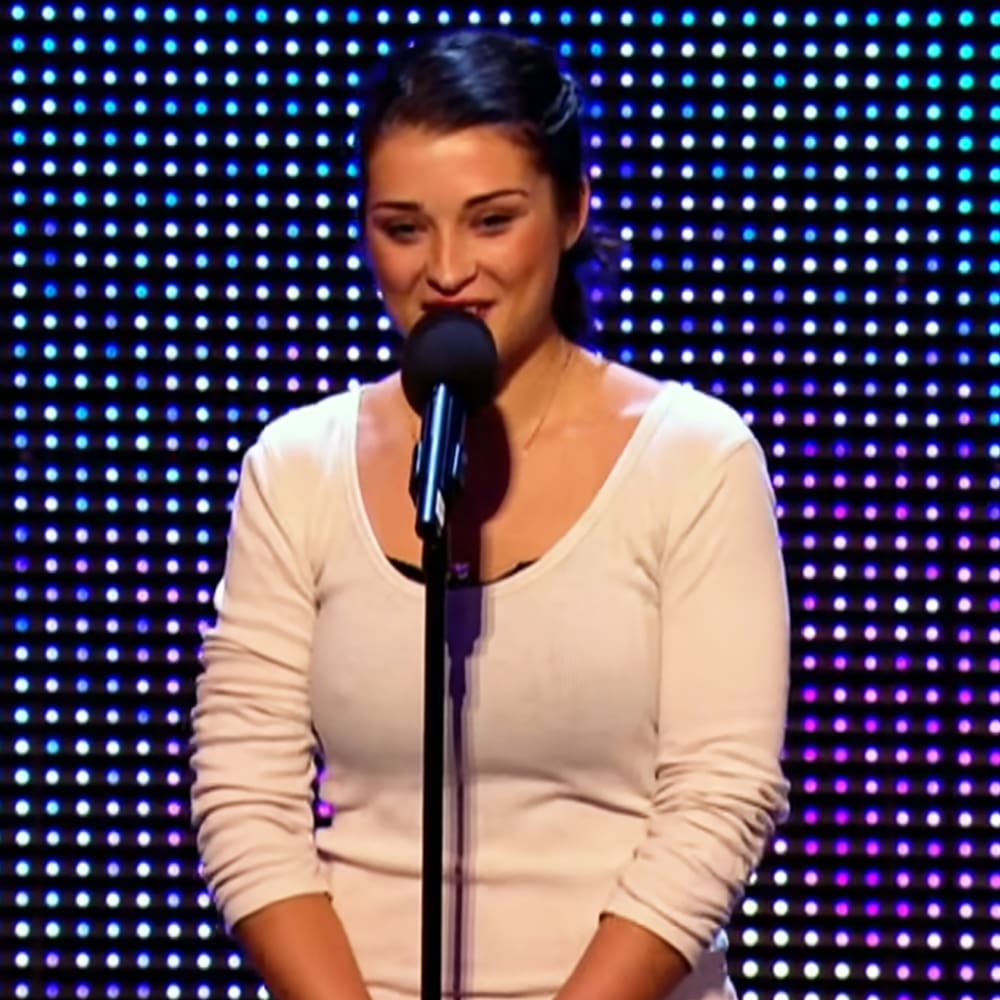 A young adult female wearing a loose, white long-sleeved top stands behind a microphone stand in an indoor performance setting with blue and purple lit-up dots on the backdrop.